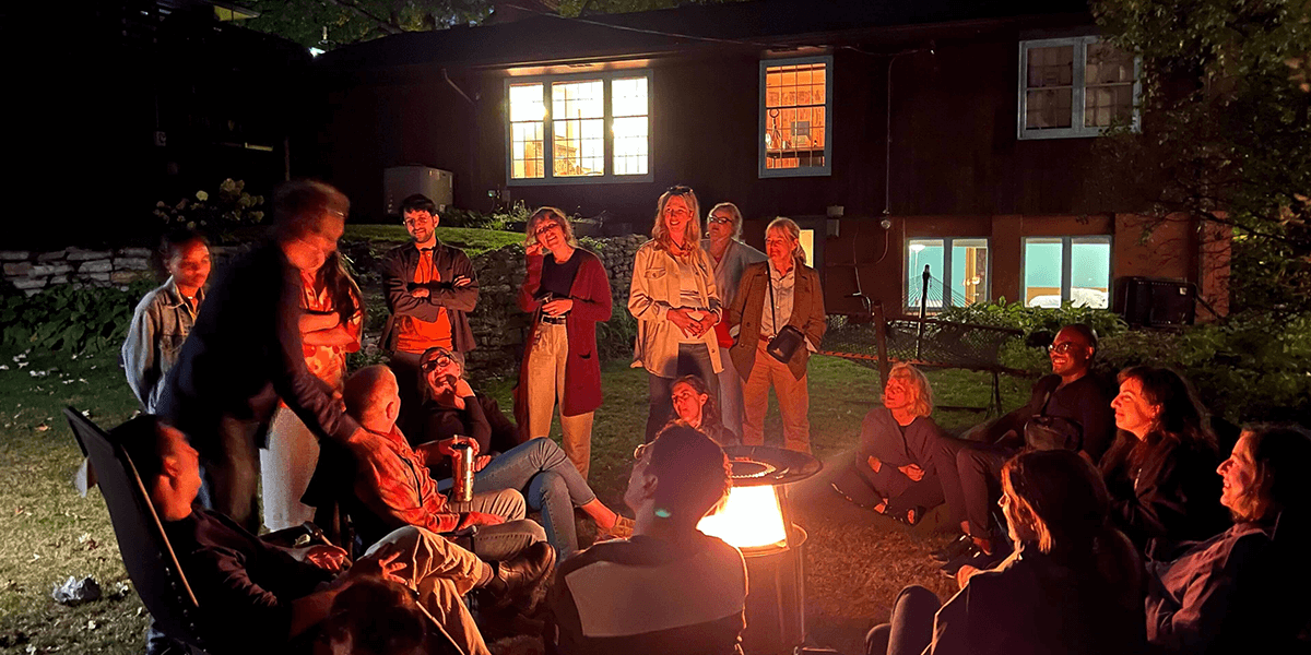 People standing and sitting around a recreational fire in a backyard. A house is visible in the background.