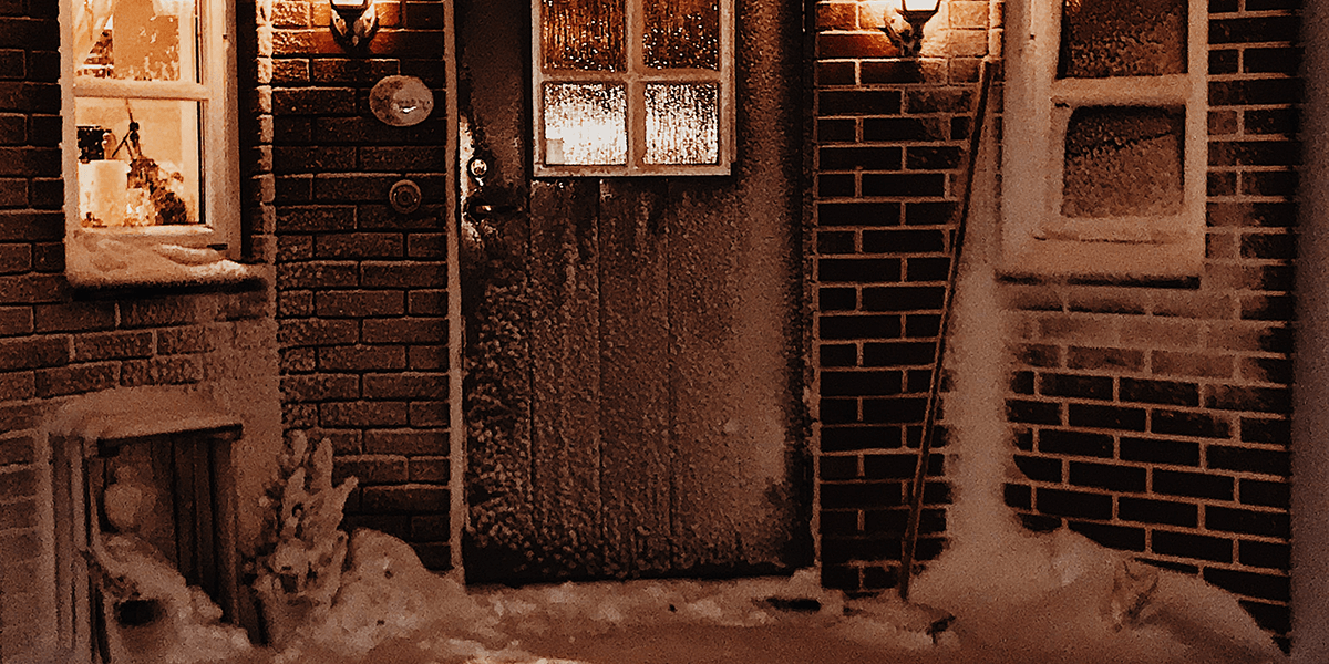 Exterior of a brick building with a window and wall light on either side of the front door. Snow is visible on the ground and on the building.