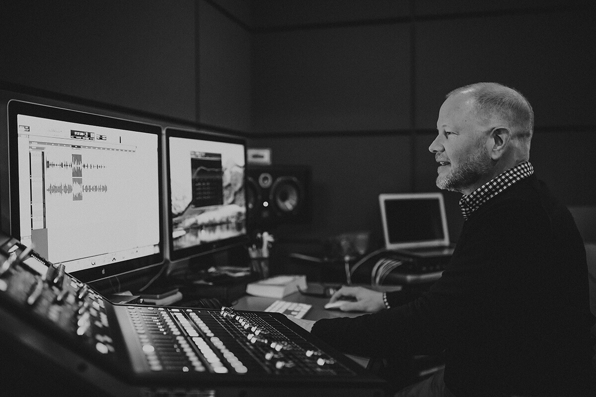 Black and white image of a man seated in front of two computer monitors on a desk. An audio mixer is visible to the left of the man.